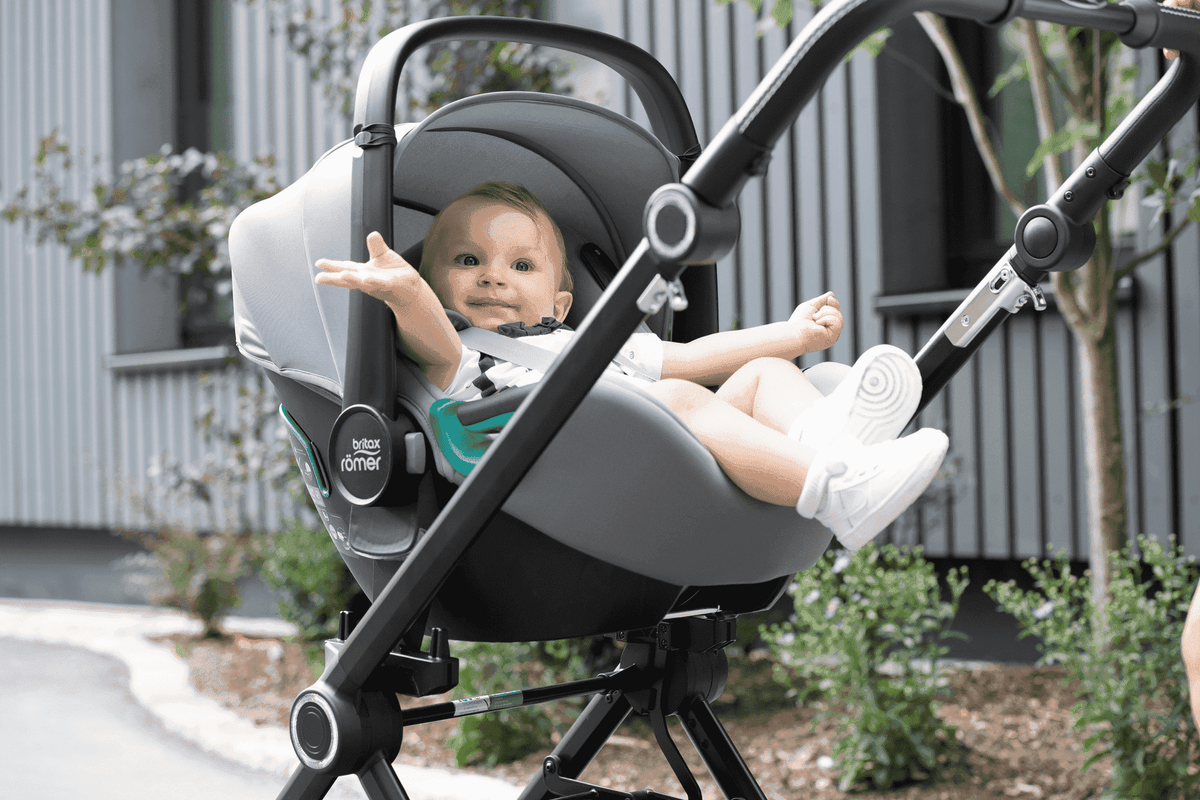 Lachendes Baby im Britax Römer Kindersitz auf Kinderwagen­gestell, weiße Schuhe, graue Schale, draußen vor modernem Gebäude