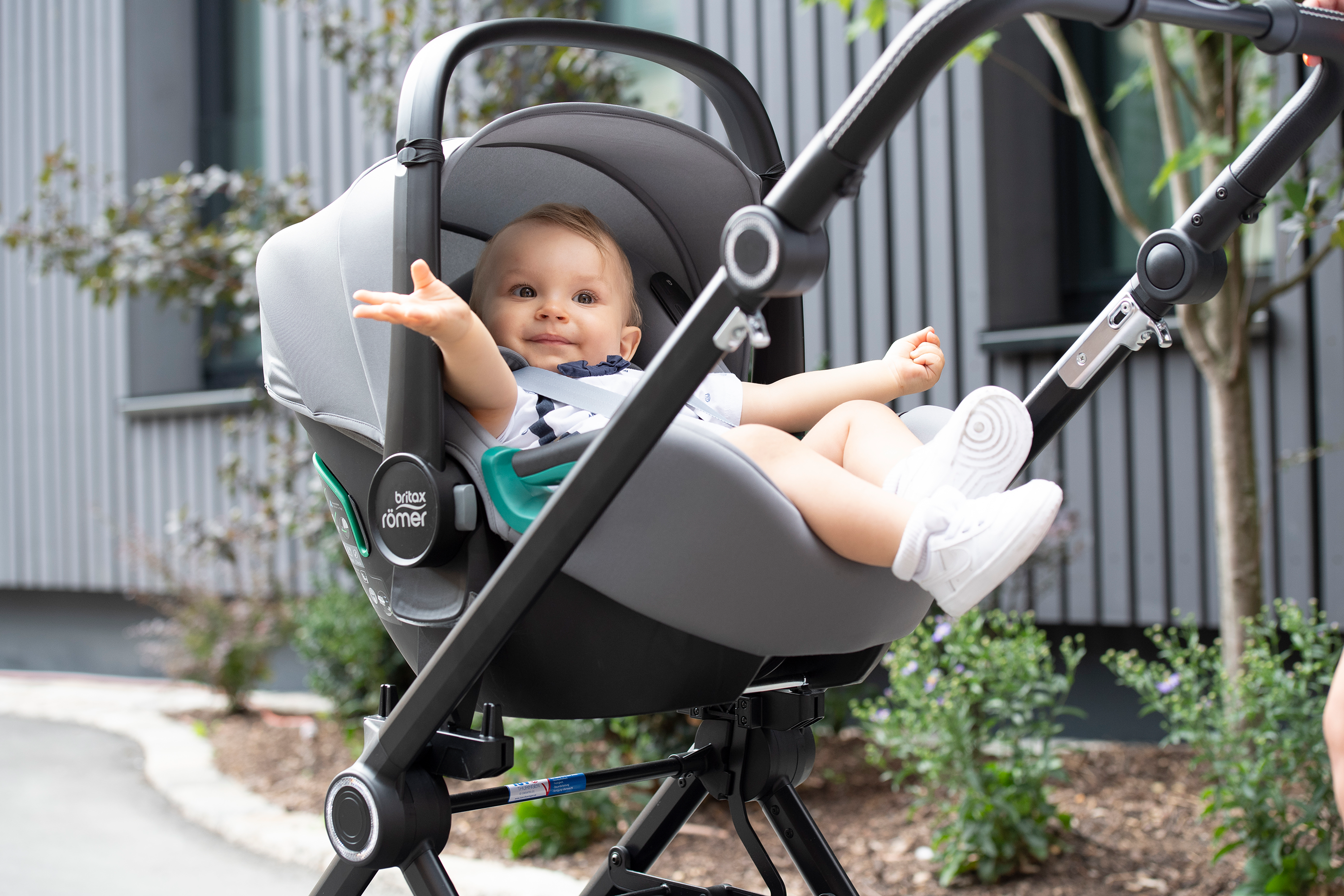 Lachendes Baby im Britax Römer Kindersitz auf Kinderwagen­gestell, weiße Schuhe, graue Schale, draußen vor modernem Gebäude