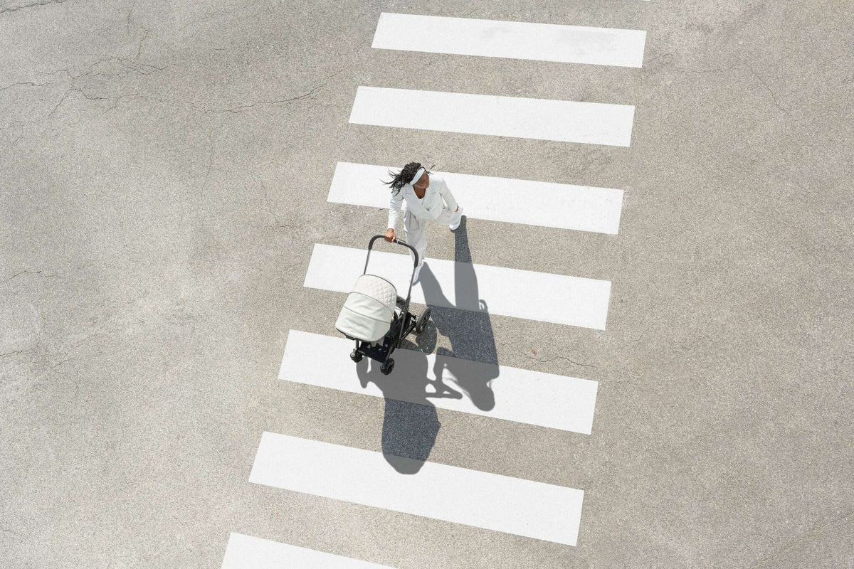 Frau in weißem Outfit schiebt Kinderwagen über Zebrastreifen von oben gesehen, lange Schatten auf grauem Asphalt
