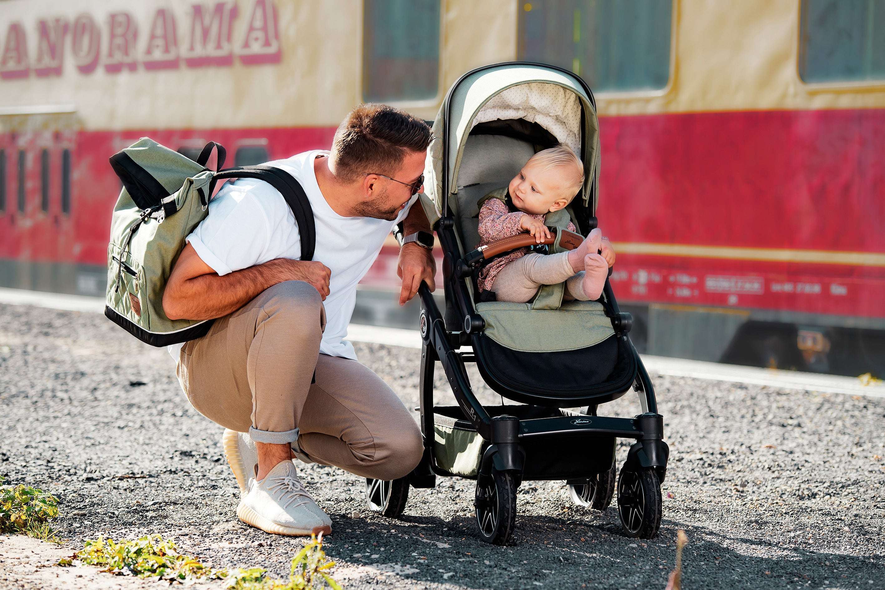 Vater hockt neben Kinderwagen, spricht mit Baby, trägt grünen Rucksack, Sandalen, weiße T‑Shirt, Baby sitzt entspannt
