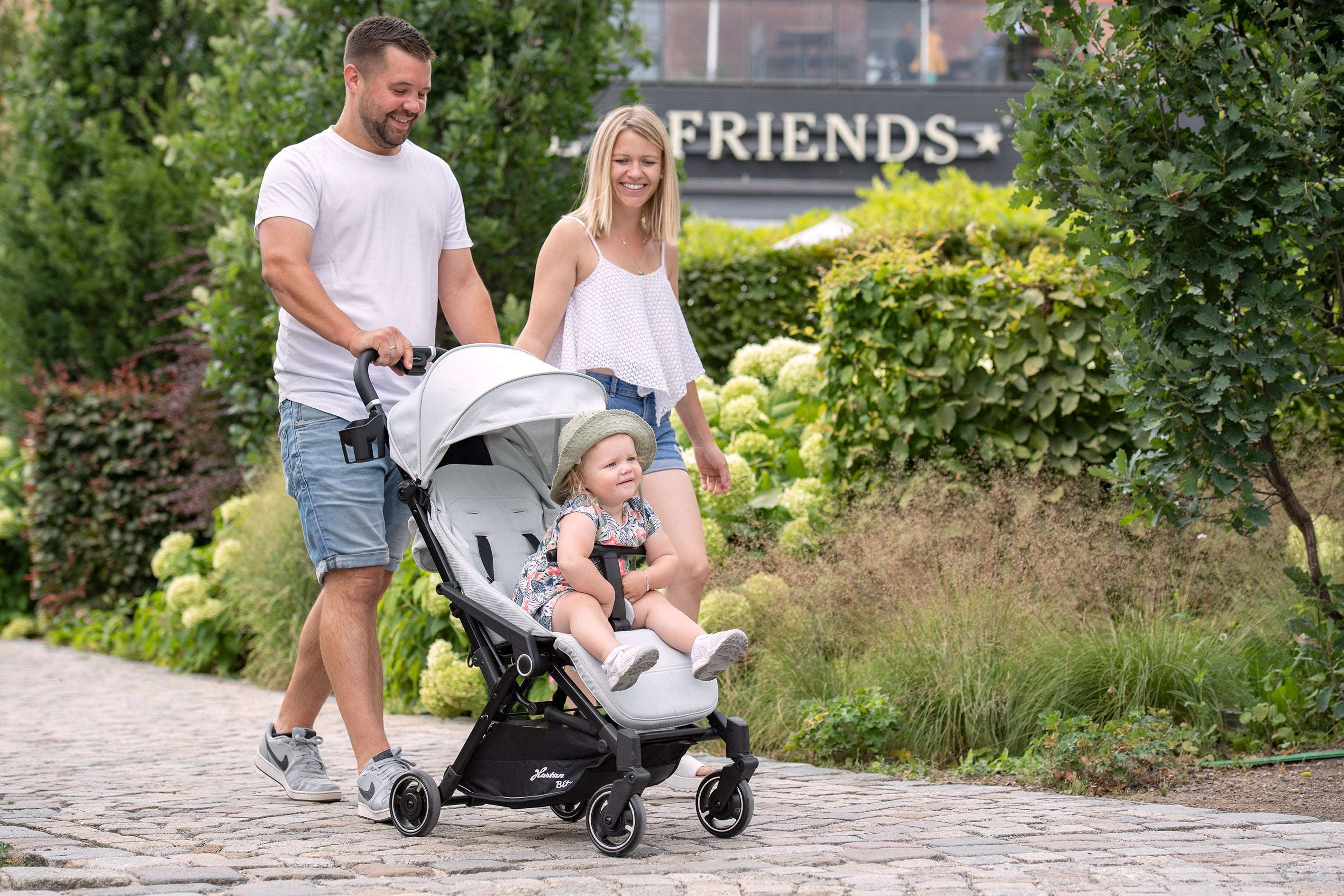 Vater und Mutter schieben Kleinkind im grauen Kinderwagen Naturkind Buggy über gepflasterten Weg in grünem Parkbereich